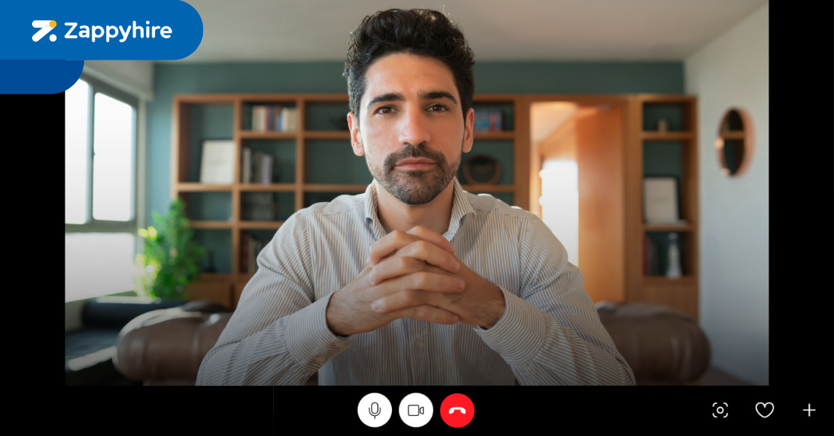 Screenshot of a one-way video interview interface with a professional man sitting in a well-lit, organized room, ready for the interview. The Zappyhire logo is visible at the top left corner of the interface, with controls for microphone, video, and call at the bottom of the screen.